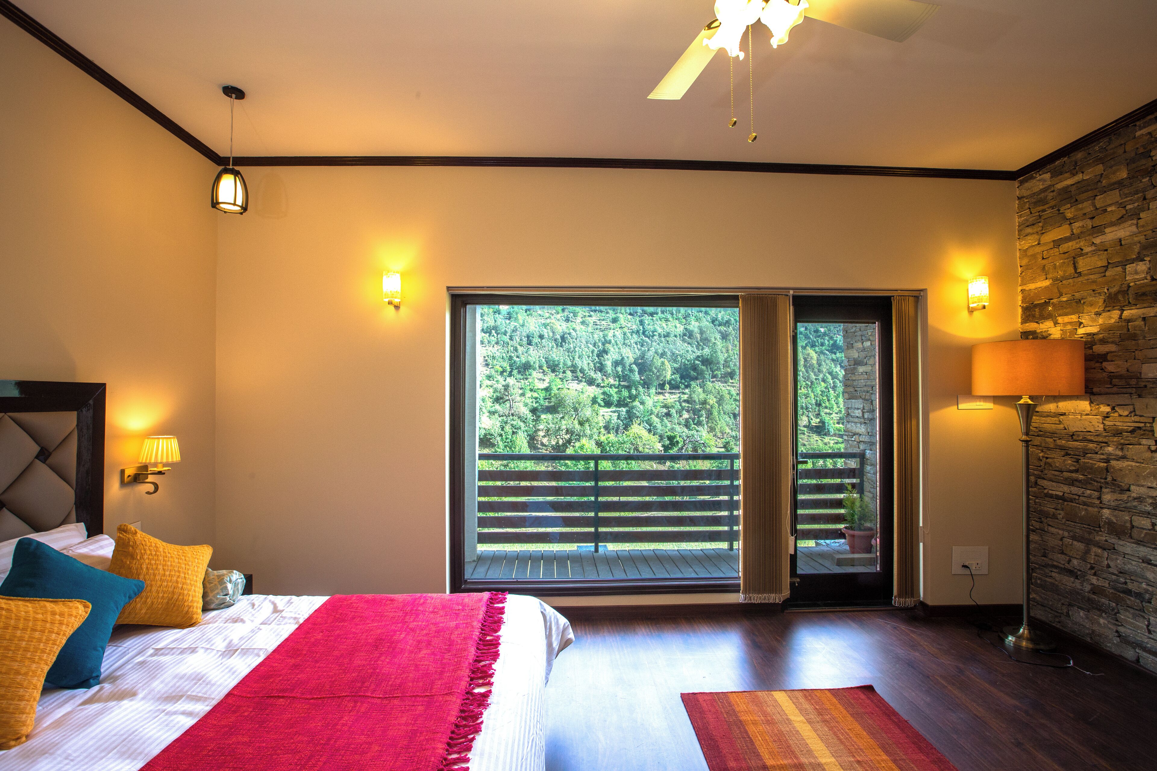 Un dormitorio con una cama grande, piso de madera, balcón con vistas a la vegetación y una pared de piedra.