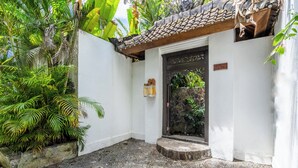 Une cour avec une entrée surmontée d’un toit en tuiles, une porte en pierre et une végétation luxuriante.