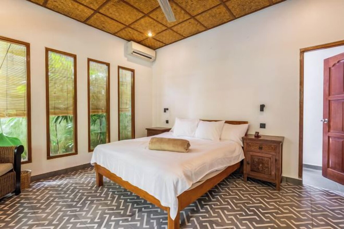 Una camera da letto con un letto grande, una testata imbottita in legno e un pavimento decorato. Si vedono tre finestre con vista su piante e un'uscita sulla destra.