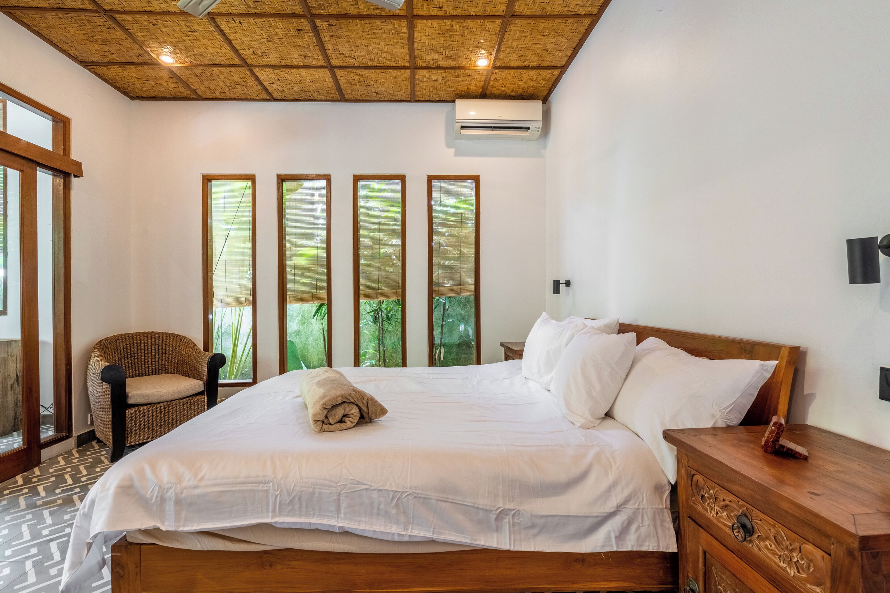Una camera da letto con un letto grande, un comodino in legno, una sedia in vimini e una finestra con vista sul verde.