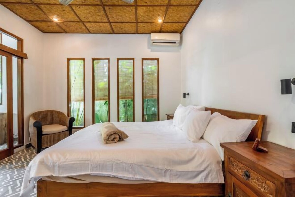 Camera da letto con un letto di legno, un comodino, una sedia e una finestra con vista sul verde.