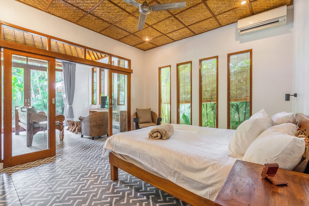 Une chambre à coucher avec un grand lit, une tête de lit en bois et une vue sur l’extérieur grâce à de grandes fenêtres.