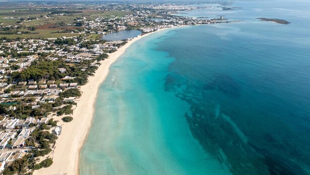 An aerial view of a coastal area with a long sandy beach, clear turquoise waters, and a densely populated residential area with houses and buildings.