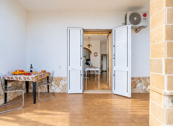 An open door leading to a dining area with a table set for two, a chair, and a table with fruits and bread.