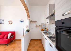 A compact kitchen with a stove, sink, and modern appliances. A red sofa is visible in the adjacent living area. - Seaside Villa Golden Shell 1 (Porto Cesareo)