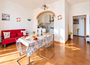 A dining area with a red sofa, a table set with plates, glasses, and a bottle, and a kitchen visible through an archway.