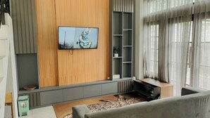 A modern living room with a flat-screen TV mounted on a wooden wall, grey shelving units, a grey sofa, and a patterned rug. - Roadsidevilla's (Kejajar)