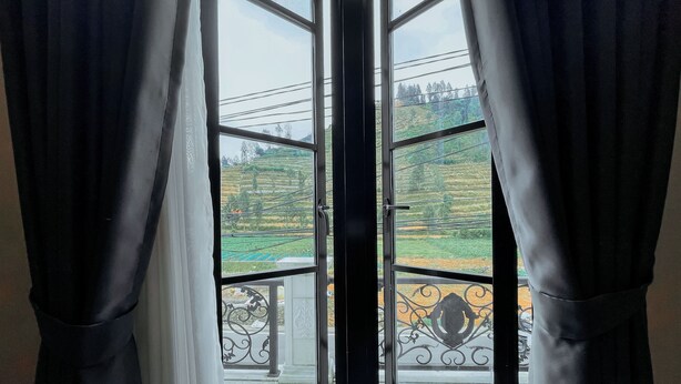 A view through a window with black curtains, revealing a balcony with a metal railing and a green landscape. - Roadsidevilla's (Kejajar)