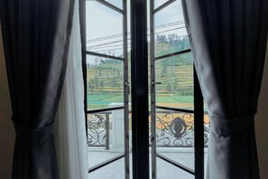 Vista a través de una ventana con cortinas negras, mostrando un balcón con barandilla metálica y un paisaje verde.