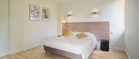 A bedroom with a bed, wooden floor, two wall-mounted lights, and two framed pictures.