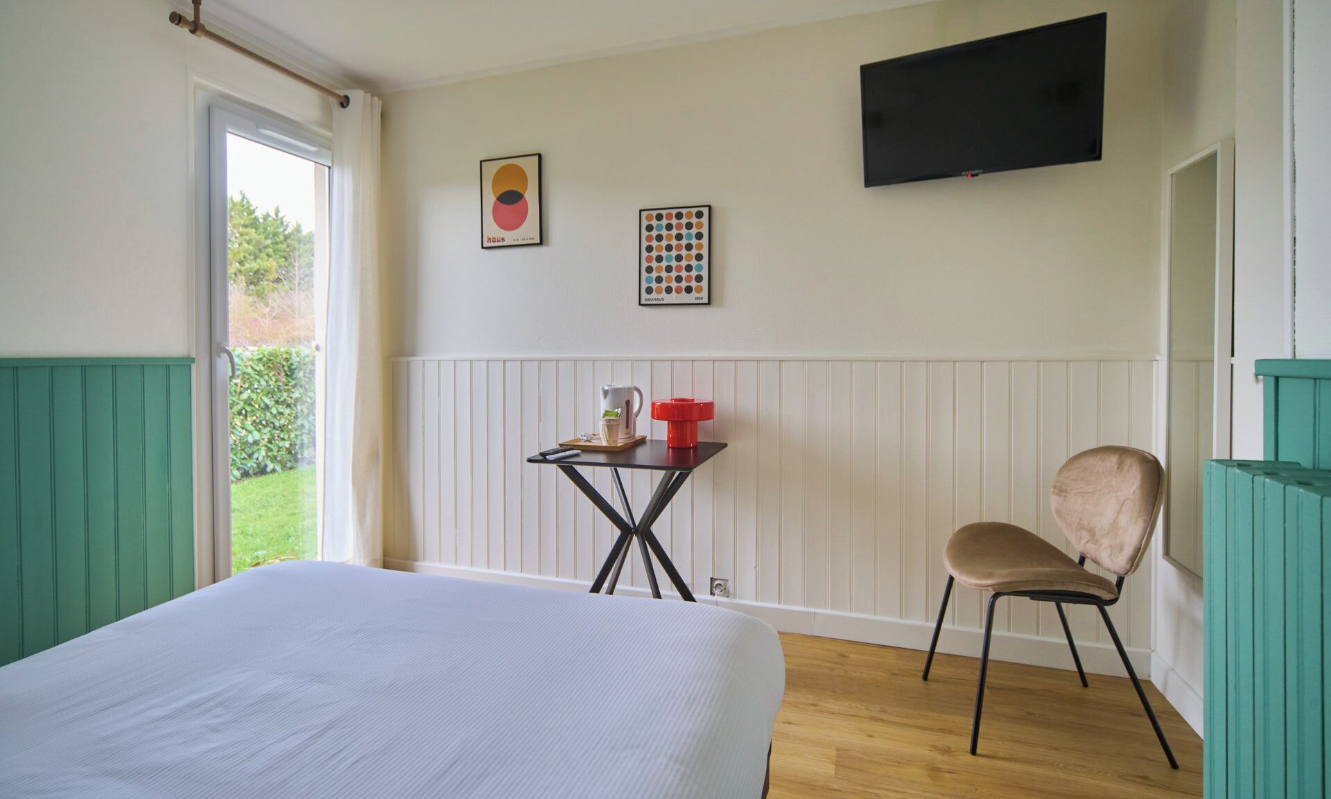 A bedroom with a bed, a chair, a small table with a kettle, and two framed pictures on the wall.