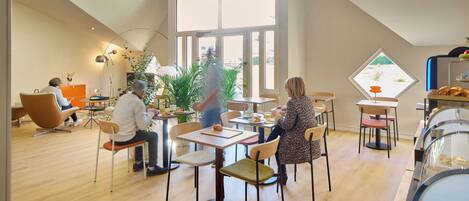 Ein moderner Essbereich mit hohen Decken und großen Fenstern, mit Holztischen und -stühlen sowie einem Buffet mit Gebäck.
