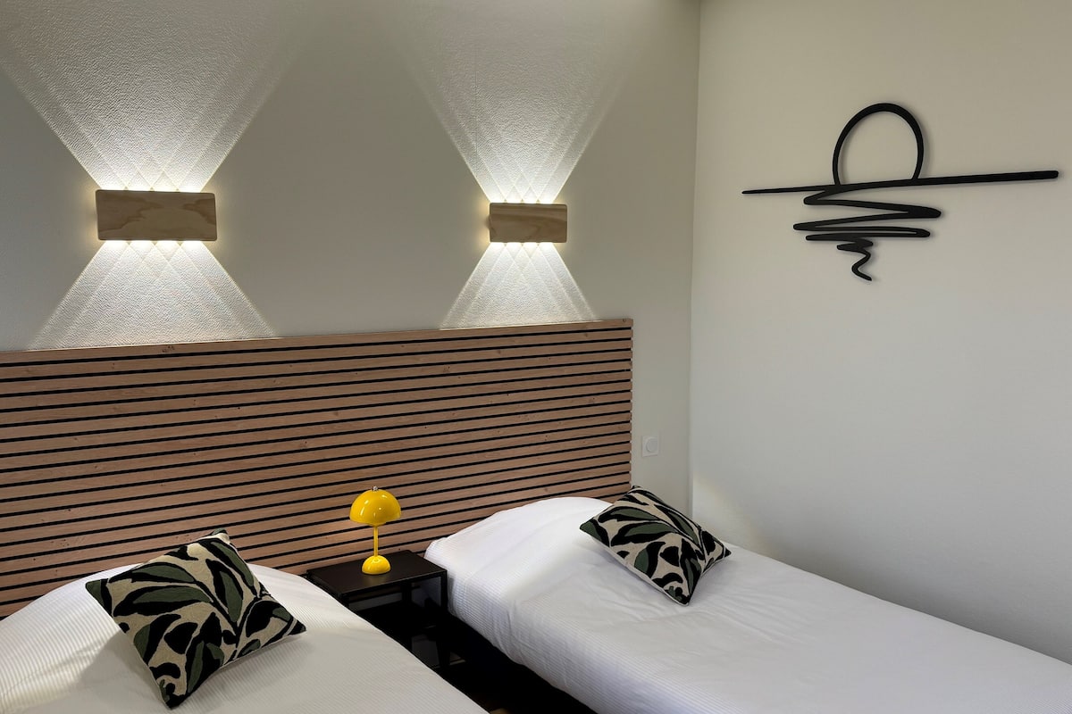 A hotel room with two single beds, a wooden headboard, a wall-mounted light fixture, and a simple abstract art piece.