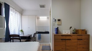 A hotel room with a bed, a wooden dresser, a desk with a chair, a small kitchen area, and a potted plant. - CraftFlat Komagome 604 (豊島区)
