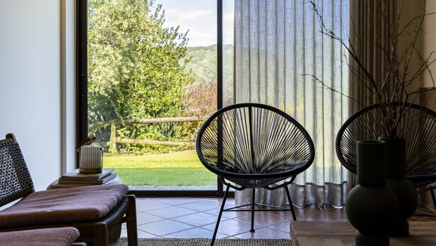 A room with a large sliding glass door leading to a garden, two black wire chairs, a wooden bench, and a vase on a table. - Ge House by House and People (Caniçada)