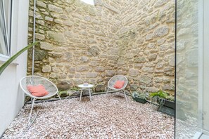 Ein Innenhof mit Steinwänden, zwei weißen Korbstühle, davon einer mit rotem Kissen, und ein kleiner runder Tisch mit einem pinken Kissen.