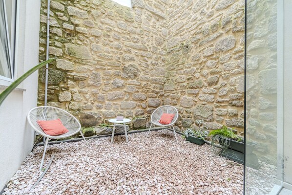 A stone-walled courtyard with two white wicker chairs, one with a red cushion, and a small round table with a pink cushion.