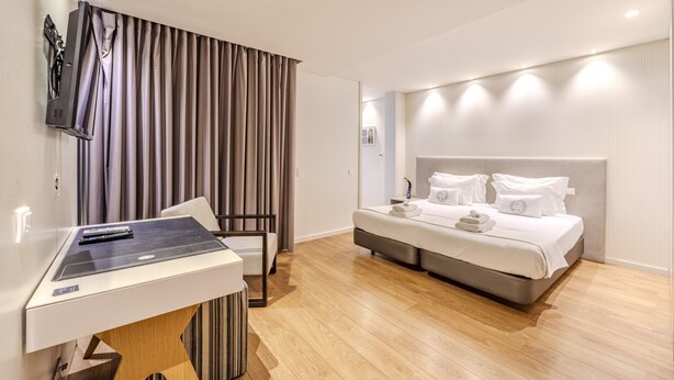Ein modernes Hotelzimmer mit Holzboden, einem Bett mit weißer Bettwäsche, einem Schreibtisch mit Computer und einem an der Wand befestigten Fernseher.