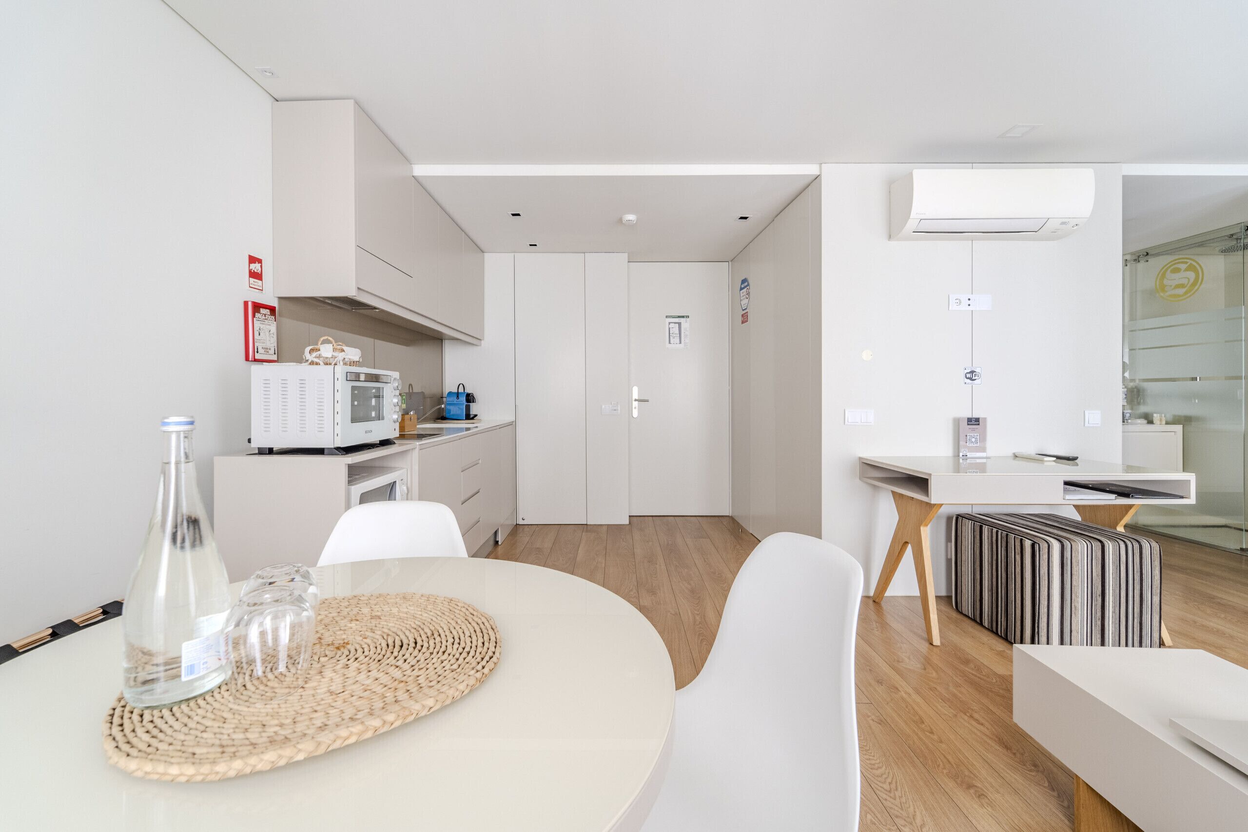 A modern kitchen with white cabinets, a microwave, and a coffee machine. There is a dining area with a round table and chairs, and a small kitchenette with a fridge and a microwave.