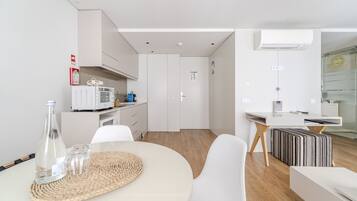 A modern kitchen with white cabinets, a microwave, and a coffee machine. There is a dining area with a round table and chairs, and a small kitchenette with a fridge and a microwave.