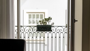 Un balcón con barandilla de hierro forjado, una planta en maceta y un edificio blanco al fondo.