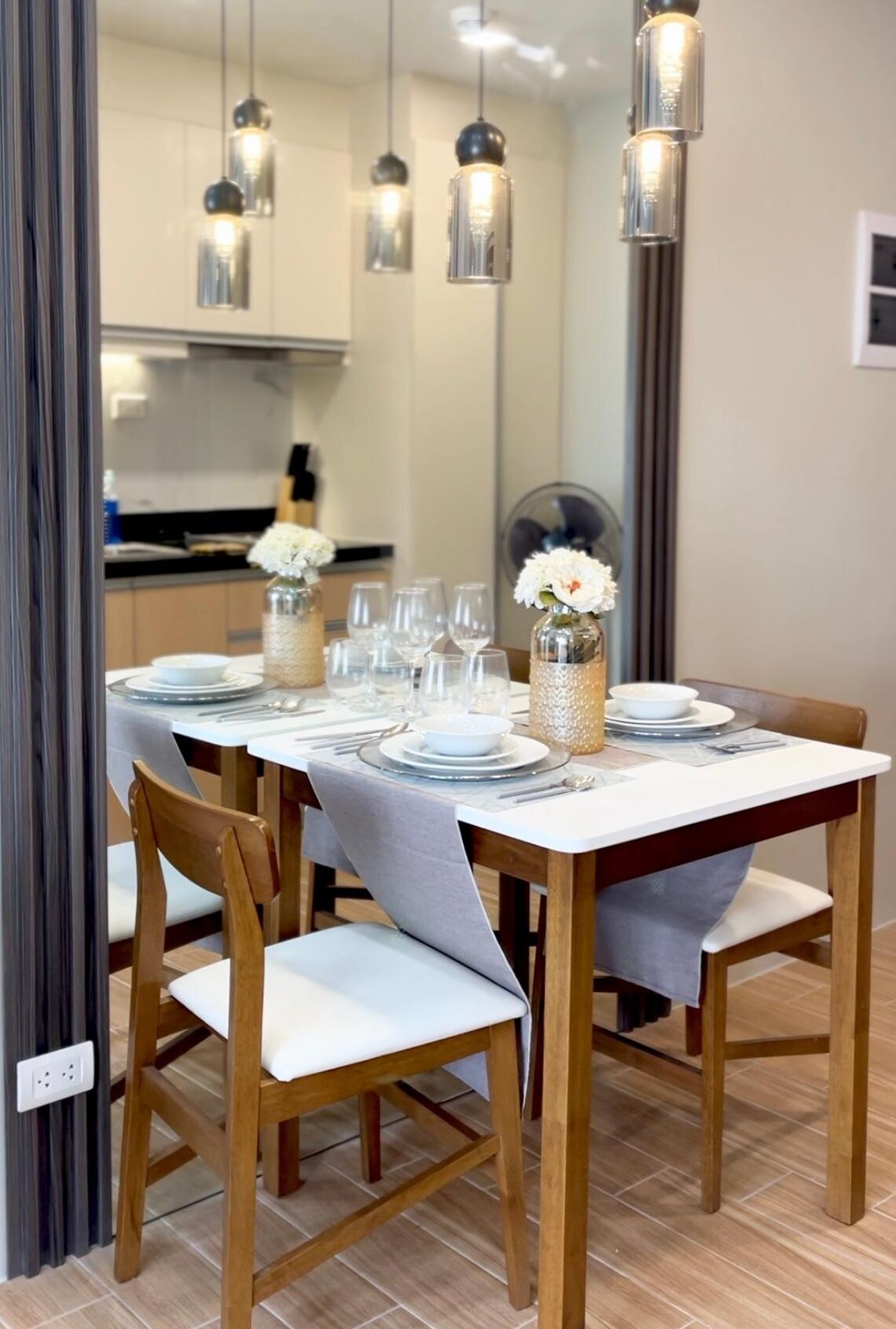 A dining area with a table set for four, wooden chairs, hanging pendant lights, and a vase with flowers.