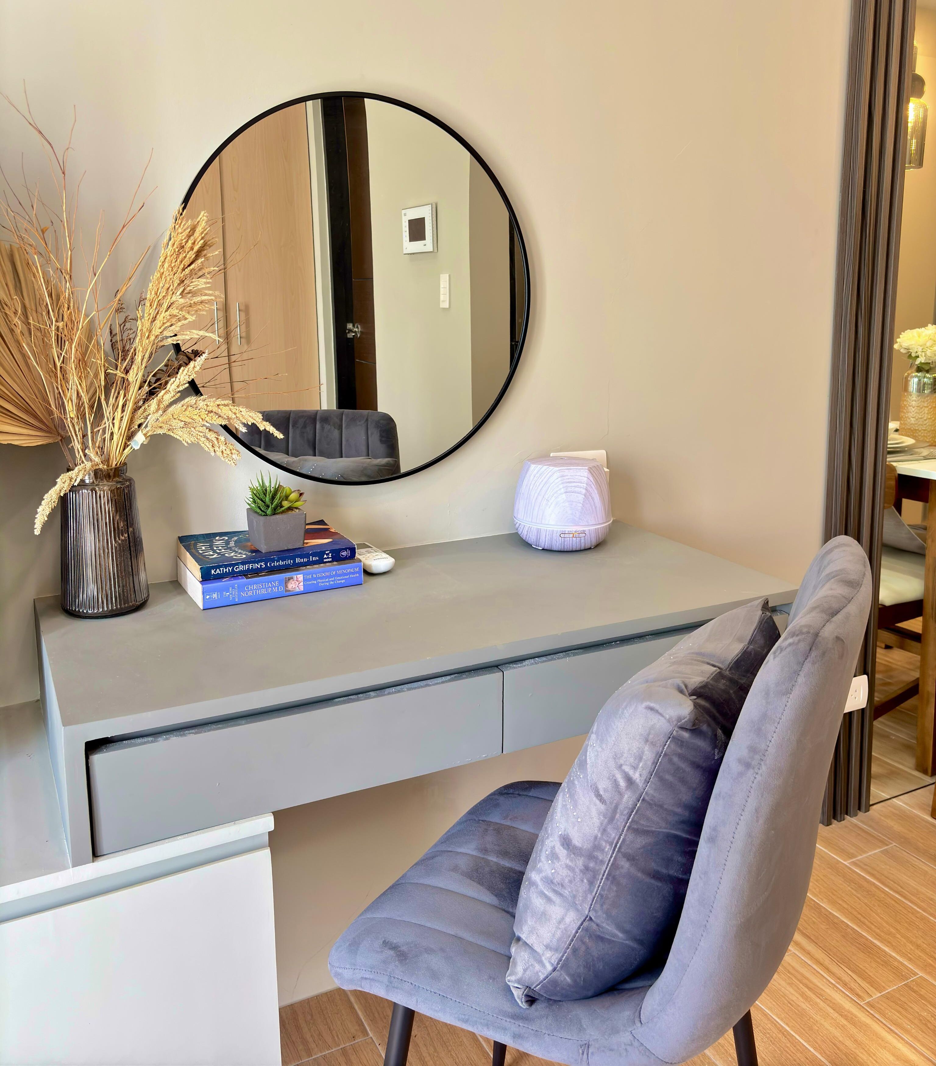 A modern room with a grey desk, a round mirror, a grey chair with a cushion, a vase with dried plants, and a small potted plant.
