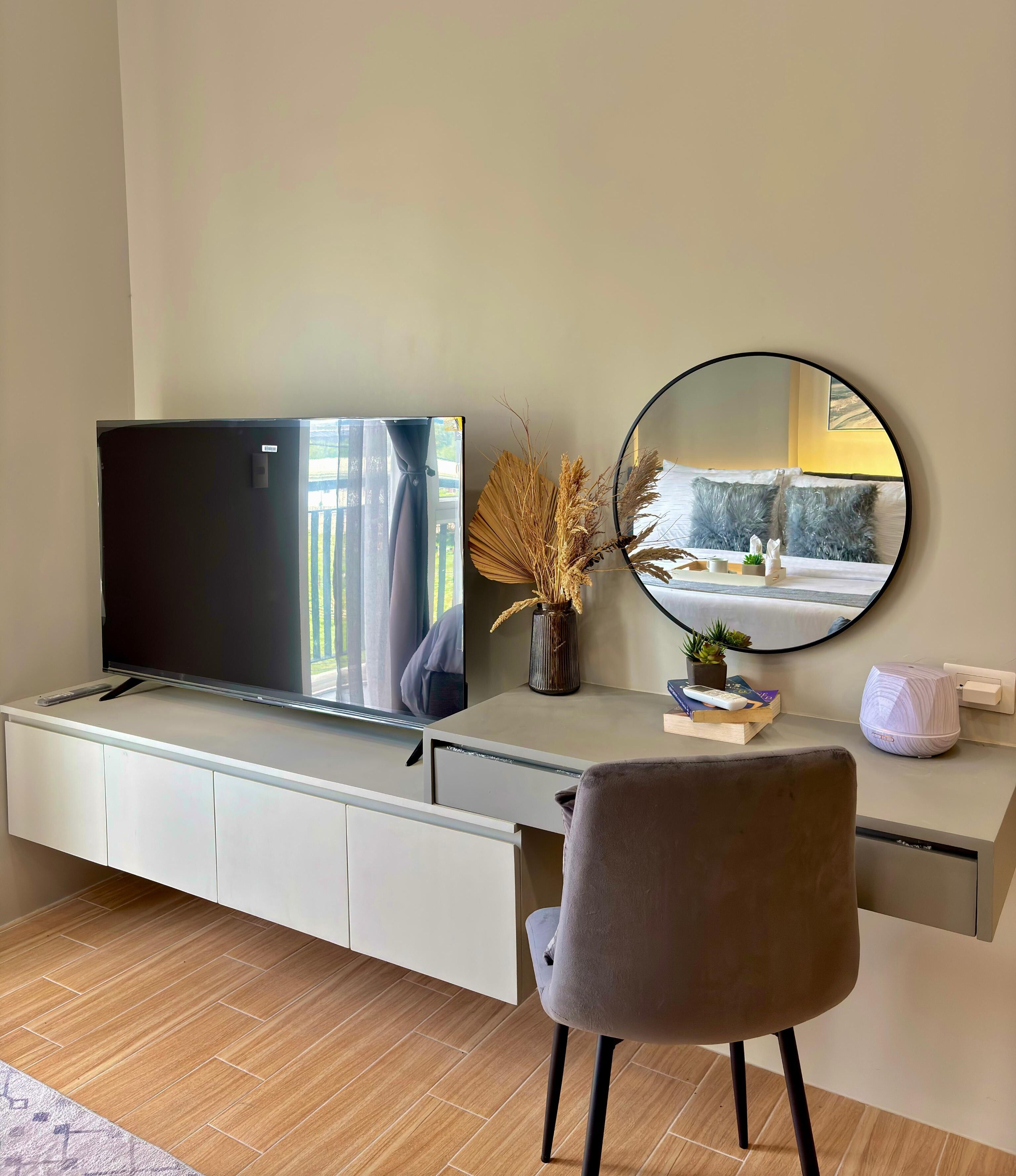 A modern room with a flat-screen TV, a round mirror, a grey chair, and a white desk.