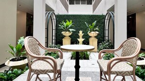 Un patio con dos sillas de mimbre y una mesa, rodeado de plantas en macetas y una pared verde.