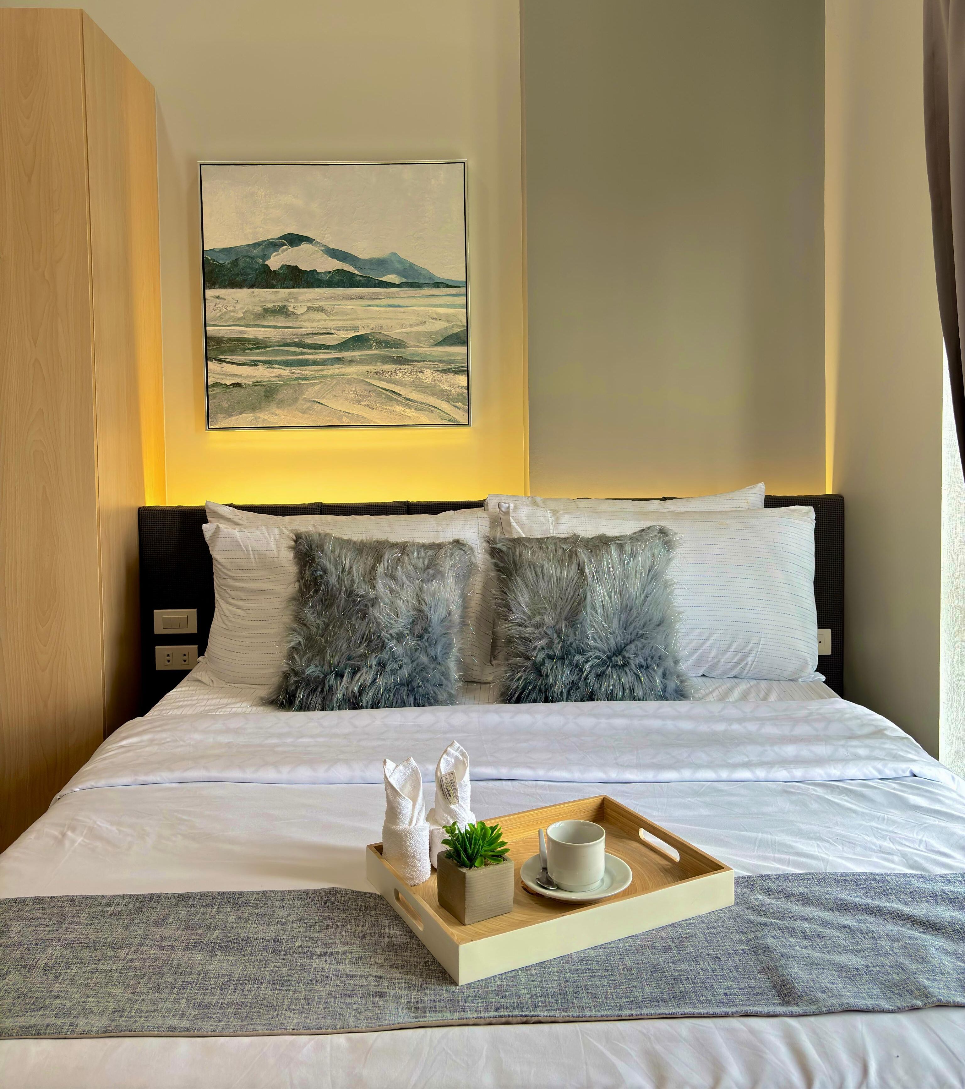 Una cama bien hecha con ropa blanca, una bandeja con una planta y una taza, y un cuadro de un paisaje montañoso en la pared.