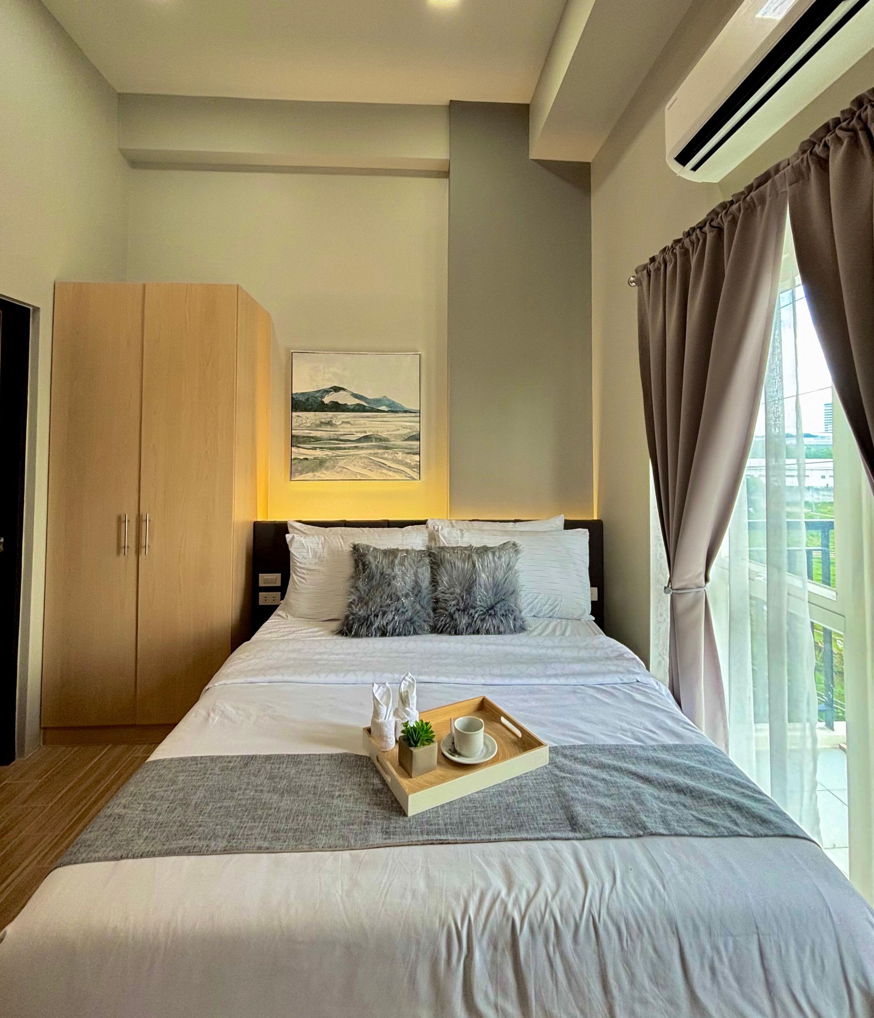 A neatly made bed with a tray of amenities, a painting on the wall, and a wardrobe.