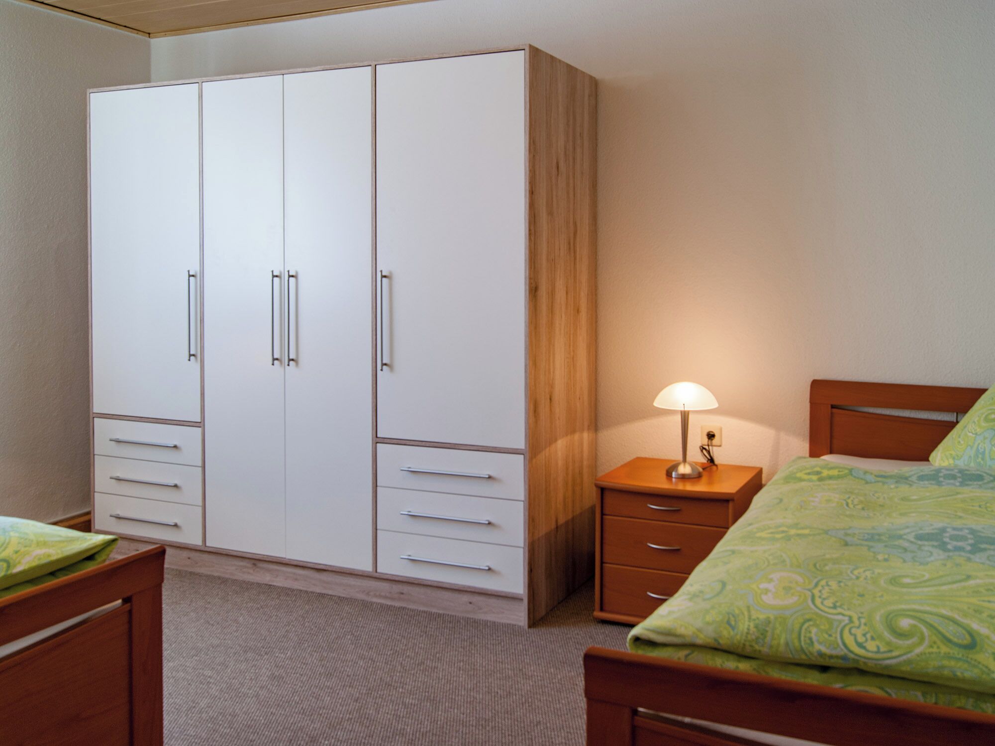 Un dormitorio con cama de madera, una mesita de noche con lámpara, un armario con varios cajones y suelo alfombrado.