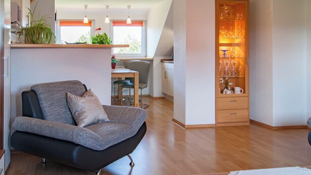 Una sala de estar moderna con un sofá gris, suelo de madera, un área de comedor con una mesa y sillas, y una cocina con refrigerador y armarios.