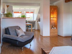 Een moderne woonkamer met een grijze bank, houten vloeren, een eethoek met een tafel en stoelen, en een keuken met een koelkast en kastjes.
