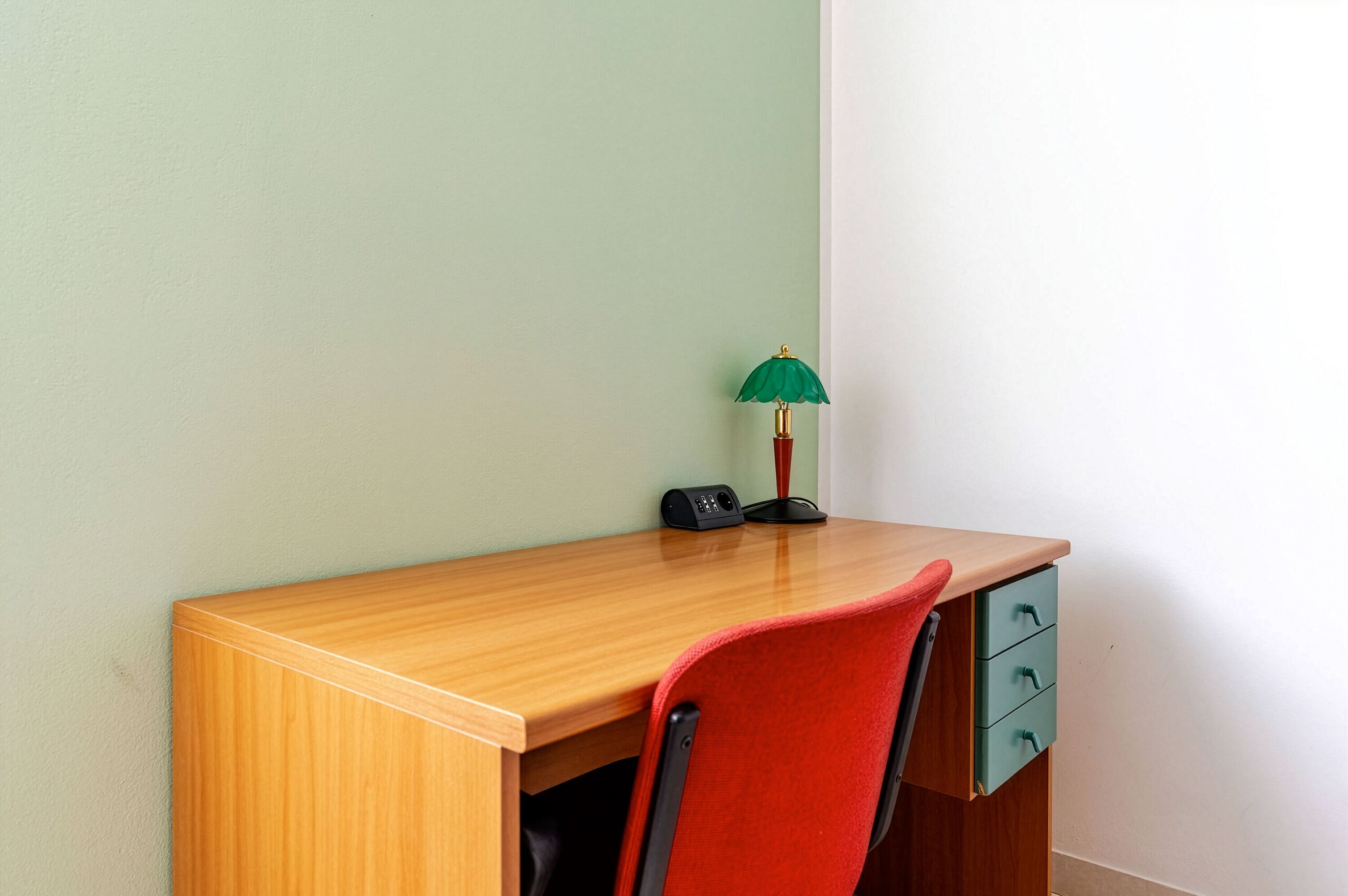 A wooden desk with a red chair, a green desk lamp, and a black speaker.