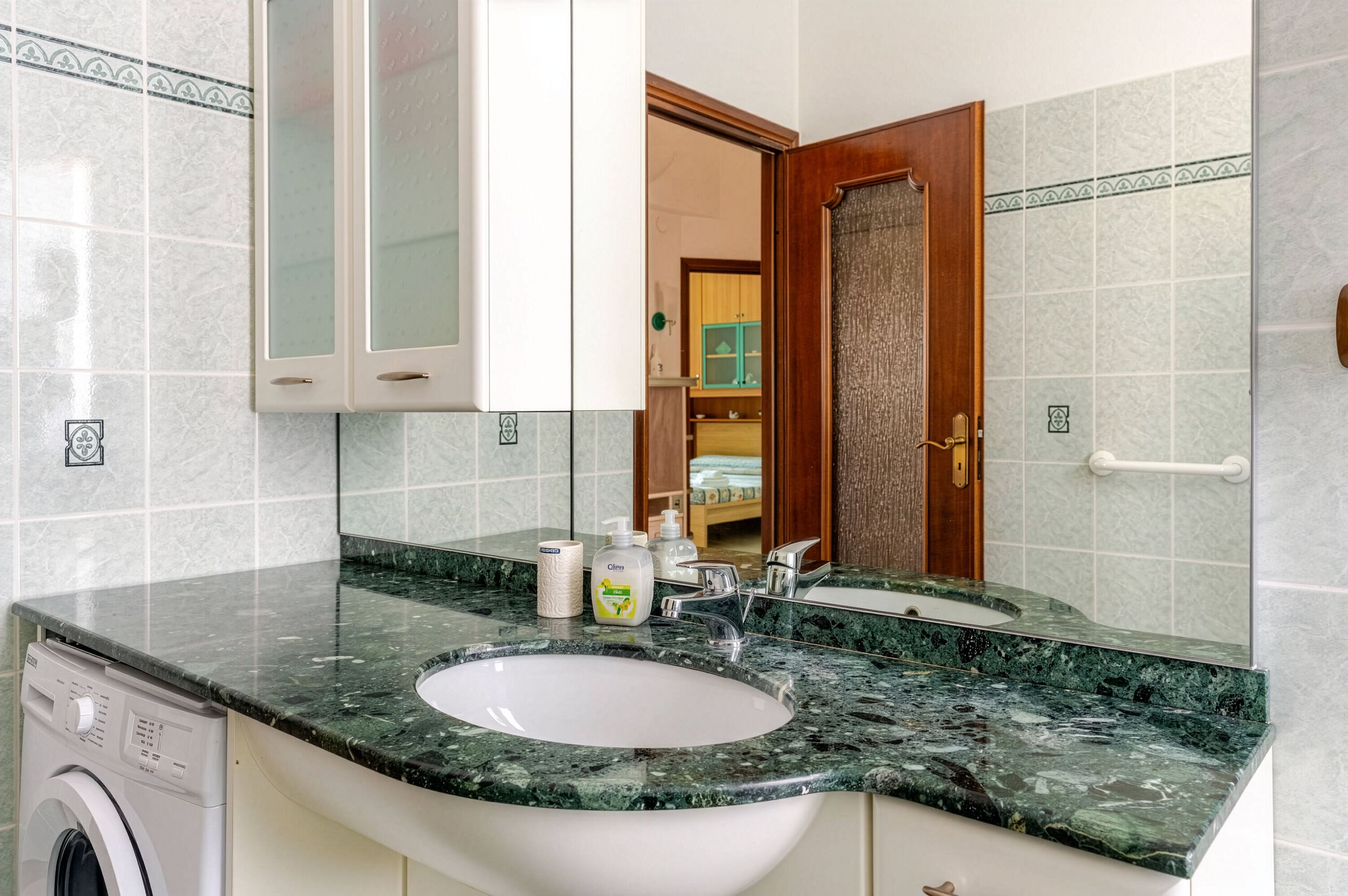 Une salle de bain avec un lavabo en marbre, un lave-linge et un miroir qui reflète une porte et une pièce où se trouve un lit.