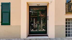 Fachada de edificio con una entrada central que presenta una reja metálica decorativa, flanqueada por dos ventanas con contraventanas verdes y una unidad de ventilación pequeña encima de la puerta.