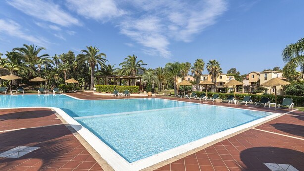 Una amplia piscina rectangular con solárium de azulejos, rodeada de palmeras y tumbonas.