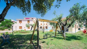 A playground with swings and a small pool area, surrounded by trees and a building with a pink facade. - Residence in Orosei with 2 bedrooms sleeps 6 (Orosei)