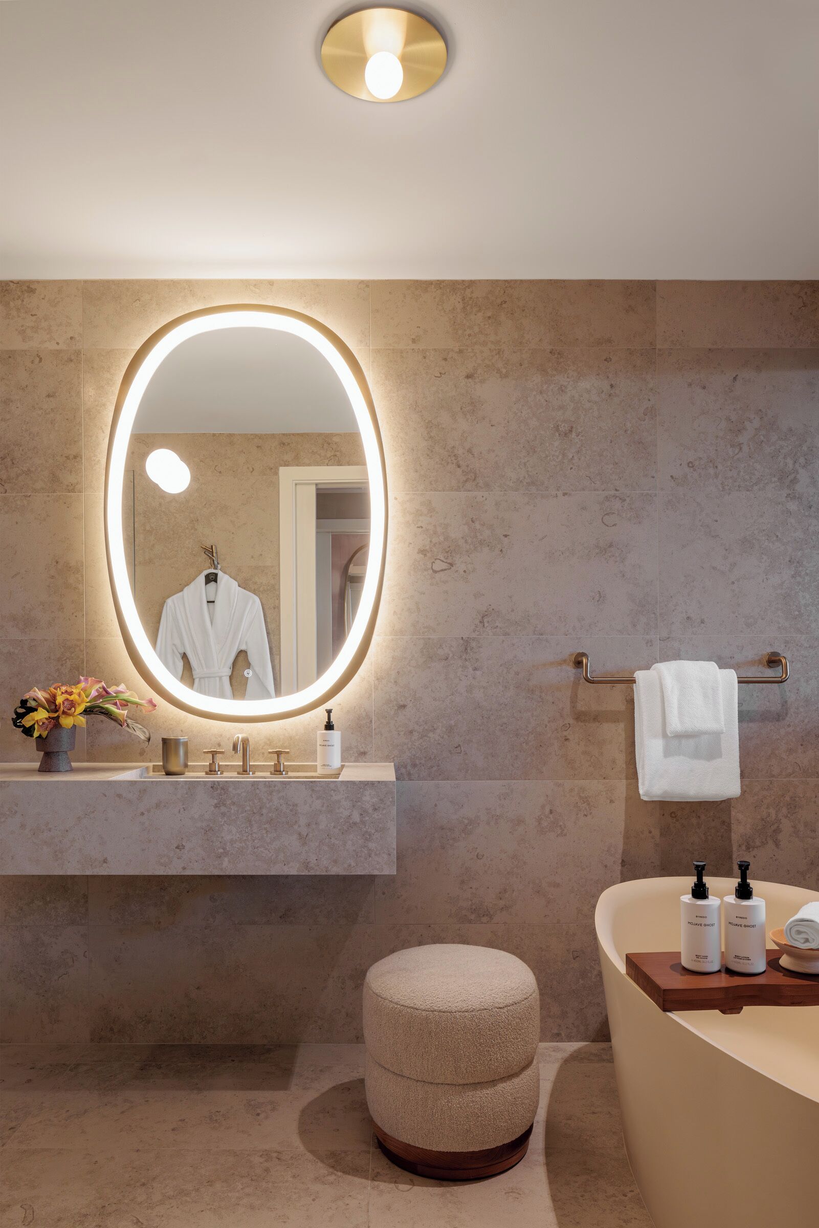 A modern bathroom with a large oval mirror, a vanity with a towel rack, and a round stool.