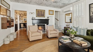 Un salón espacioso con chimenea, dos sillones, un sofá verde y una mesa de centro con libros y un jarrón de flores.