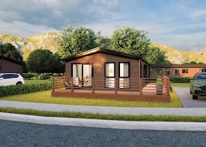A single-story wooden cabin with a deck, surrounded by greenery and mountains.