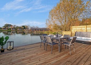Une terrasse en bois avec une table dressée pour le repas, offrant une vue sur une étendue d’eau et une rangée de bâtiments.