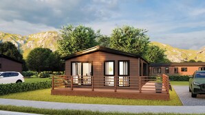 A single-story wooden cabin with a deck, surrounded by greenery and mountains. - Deluxe modern and contemporary luxury lodge perfect for a relaxing escape. (Workington)