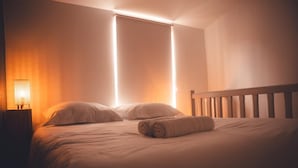 A neatly made bed with a wooden headboard, a rolled towel on the bed, and a wall-mounted light fixture.