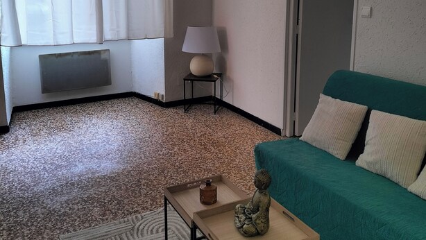 A living room with a green sofa, a small table with a decorative item, a lamp on a side table, and a window with white curtains. - Pet-friendly apartment le petit paradis :) (Saint-Pons-de-Thomières)