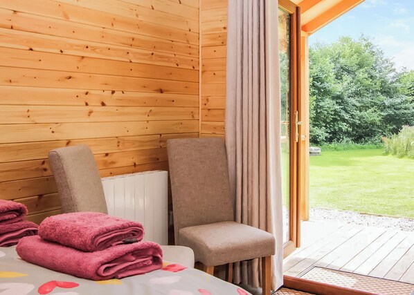 A room with wooden walls, a bed with a white headboard, two chairs, and a view of a garden through a glass door.