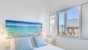 A bedroom with a bed, a painting of a beach, a window with a view of a balcony, and a nightstand. - Cosy apartment for 5 people with A/C, WIFI, TV and veranda, close to Ostuni (Pantanagianni-pezze Morelli)