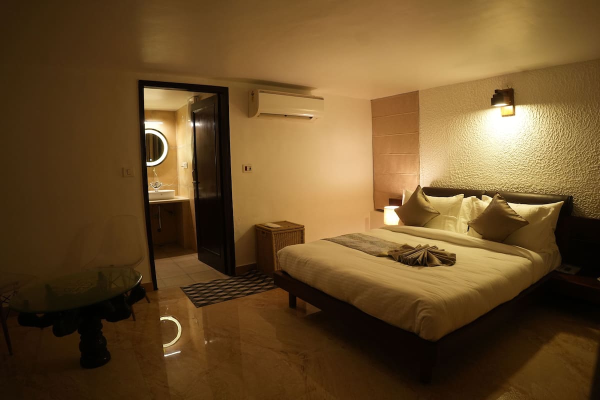 A hotel room with a bed, bedside table, lamp, and an air conditioning unit.