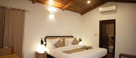 A bedroom with a wooden ceiling, a bed with white linens, a bedside table with a lamp, and a wicker chair.